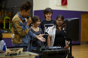 Photo of Black man with dreads showing 3 middle school students the mixing board