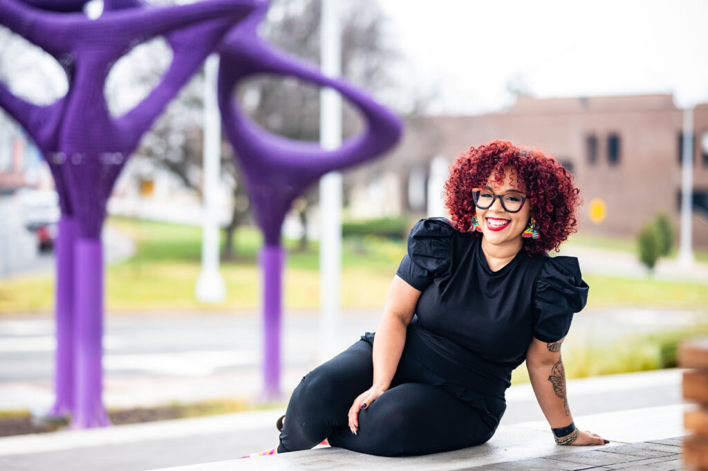 Photo of a black woman with red curly hair and glasses, sitting in front of a purple sculpture