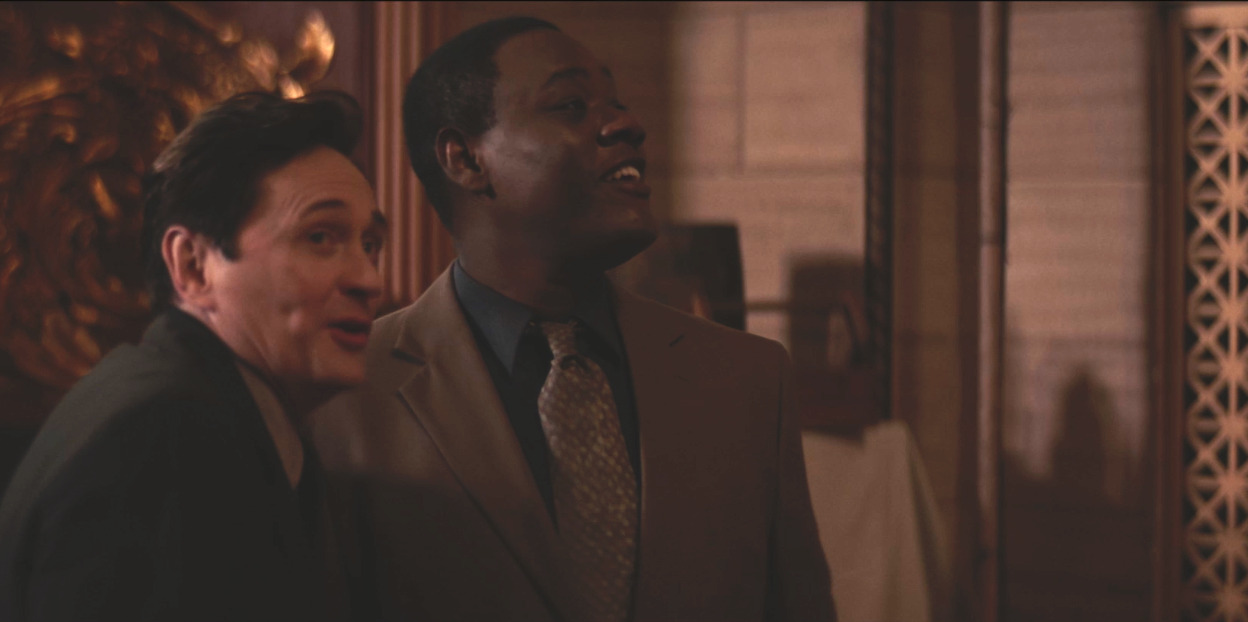 A white man in black suit and Black man in a tan suit laugh and converse