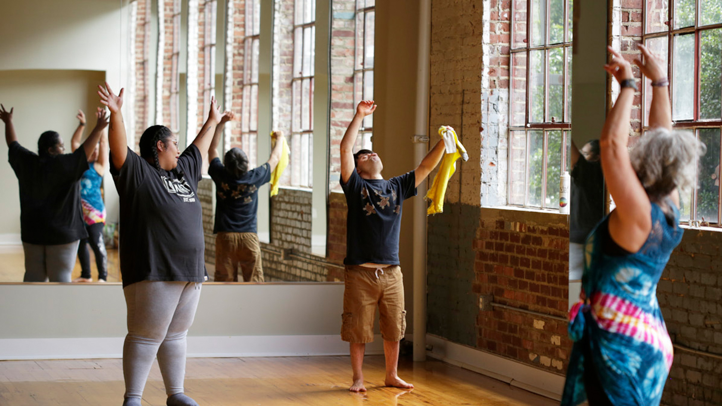 Photo of black woman, white man, and white woman all standing with their arms raised in front of a wall length mirror